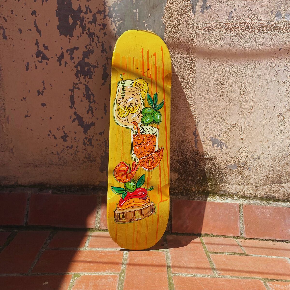 Yellow_skateboard_deck_featuring_colorful_cocktail_citrus_olive_and_pepper_illustrations_leaning_against_a_textured_wall